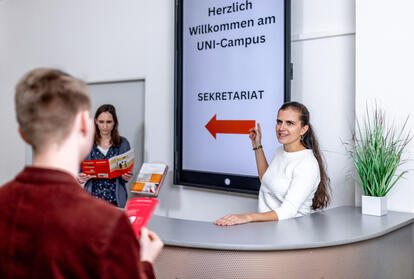 2 Personen hinter Counter, Digital Signage-Display dahinter an der Wand, ein Besucher im Vordergrund