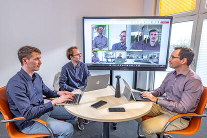 Meeting 3 Personen am Tisch mit Konferenzsystem, Teinehmer im Split-Screen im Groß-Display