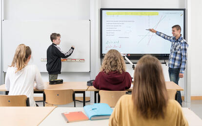 Schülerinnen und Schüler Oberstufe in Klasse mit Lehrer an Tafelkombination aus digitalem Board und klassischem Whiteboard